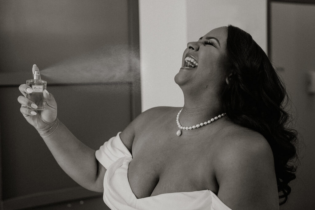 Black and white photo of a bride spraying on her perfume while laughing.