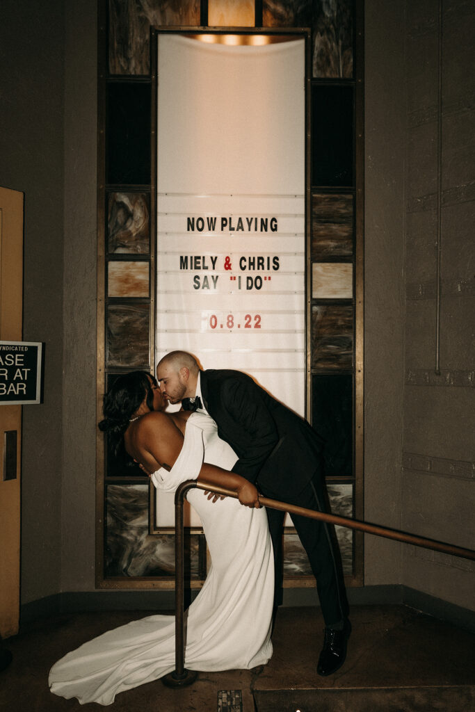 Bride and groom sharing a celebratory kiss in front of their "Now Playing" sign for their movie theatre wedding.