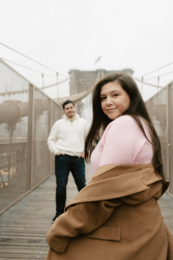 A couple walks along the Brooklyn Bridge on a cloudy day with the fog surrounding the bridge.