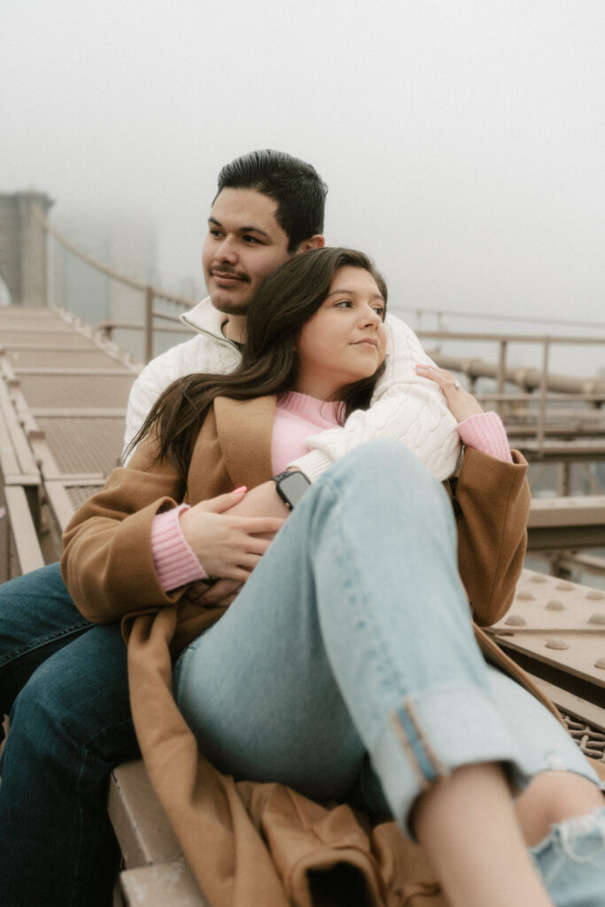 A couple cuddles together atop the side of the Brooklyn Bridge as a rainy mist surrounds them.