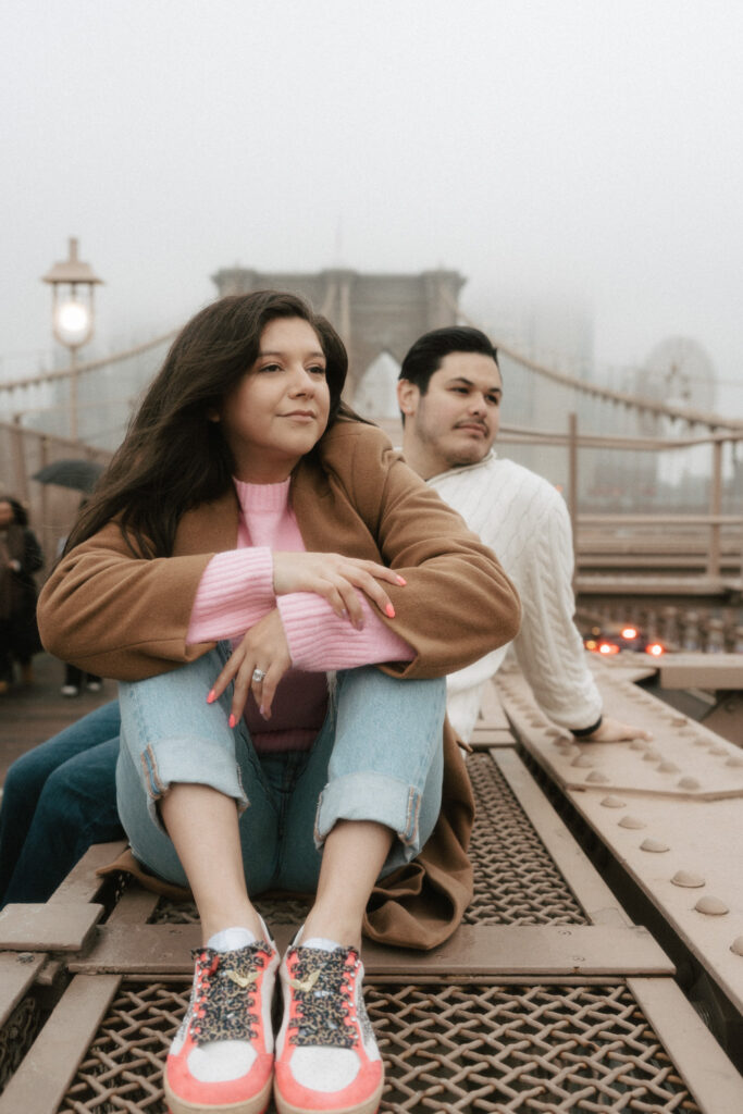 A couple sits atop the side of the Brooklyn Bridge as a rainy mist surrounds them.