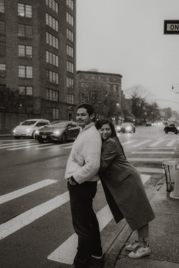 A woman snuggly leans onto her husband on a sidewalk in West Village with cars whizzing by in the background.