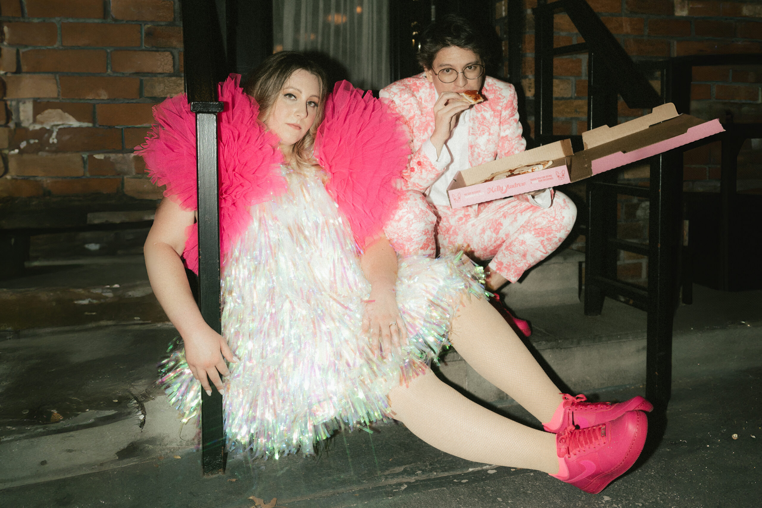 A bride in a custom sparkly disco dress with pink tulle sleeves eating pizza with her groom who's wearing a pink patterned suit.