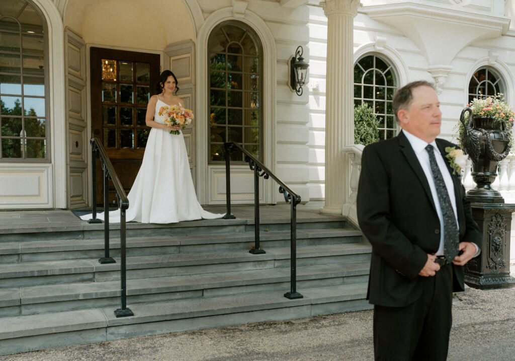 A bride approaches her father who is facing away to do a father/daughter first look on her wedding day.