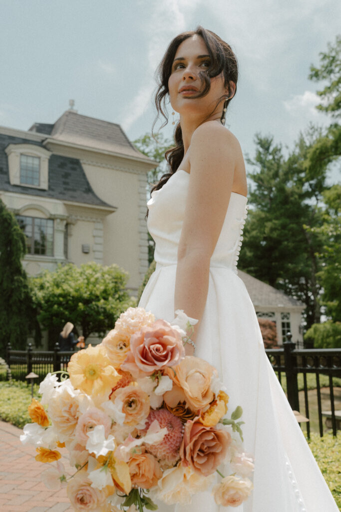 A bride stands with her flowers by her side and the wind blowing through the loose strands of her ponytail.