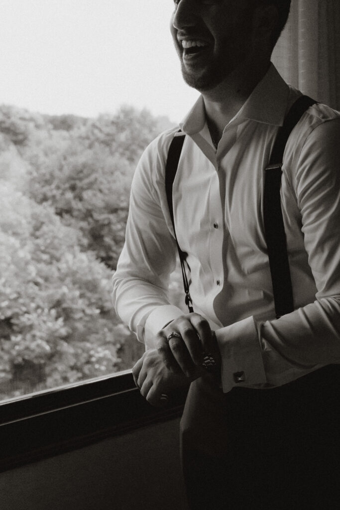 In black and white, a groom gets ready by a window for his wedding.