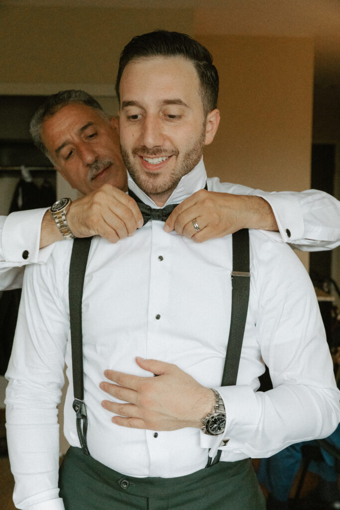 A groom's father reaches around him to straighten his bowtie.