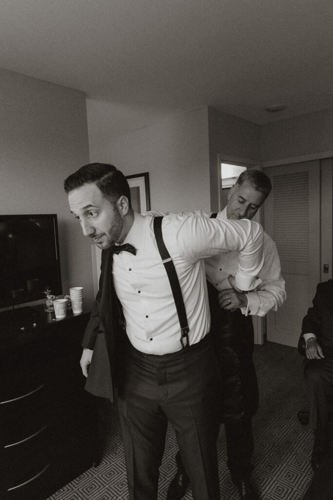 In black and white, a groom is being helped with his jacket by his father.