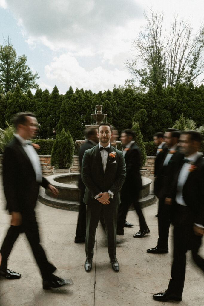 A groom stands handsomely while his groomsmen move quickly around him and are blurred from the motion.