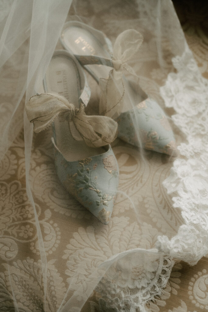 A bride's blue floral slingback heels lie on a couch underneath her sheer, lace-trimmed veil.