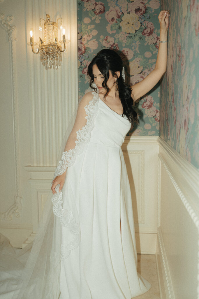 A bride leans beautifully against a blue floral wallpapered wall in the bathroom of the Ashford Estate.