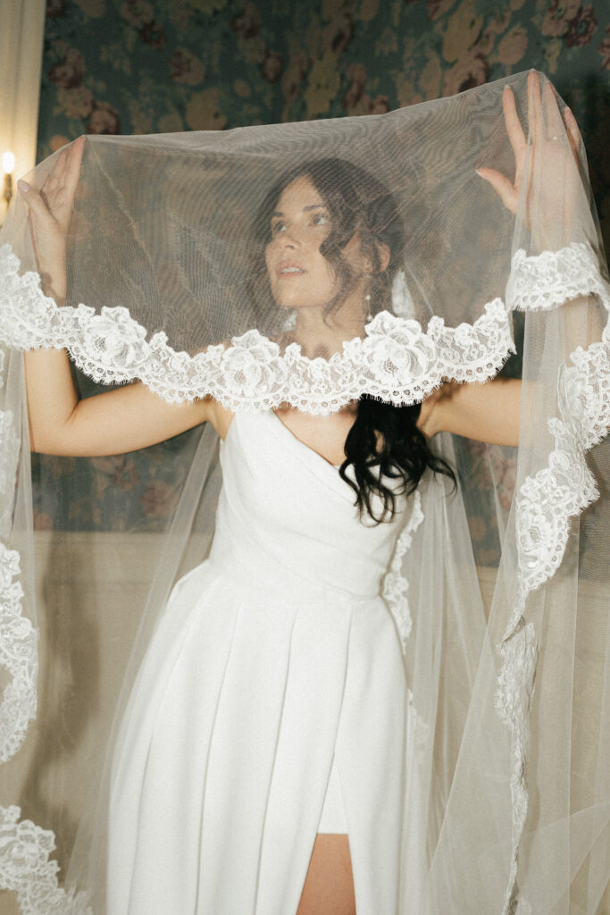 A bride's lace-trimmed veil drapes over her hands and face with blue wallpaper in the background.