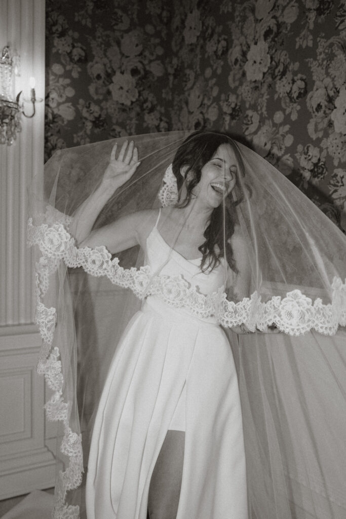 A bride playfully laughs with her veil covering her, in black and white.