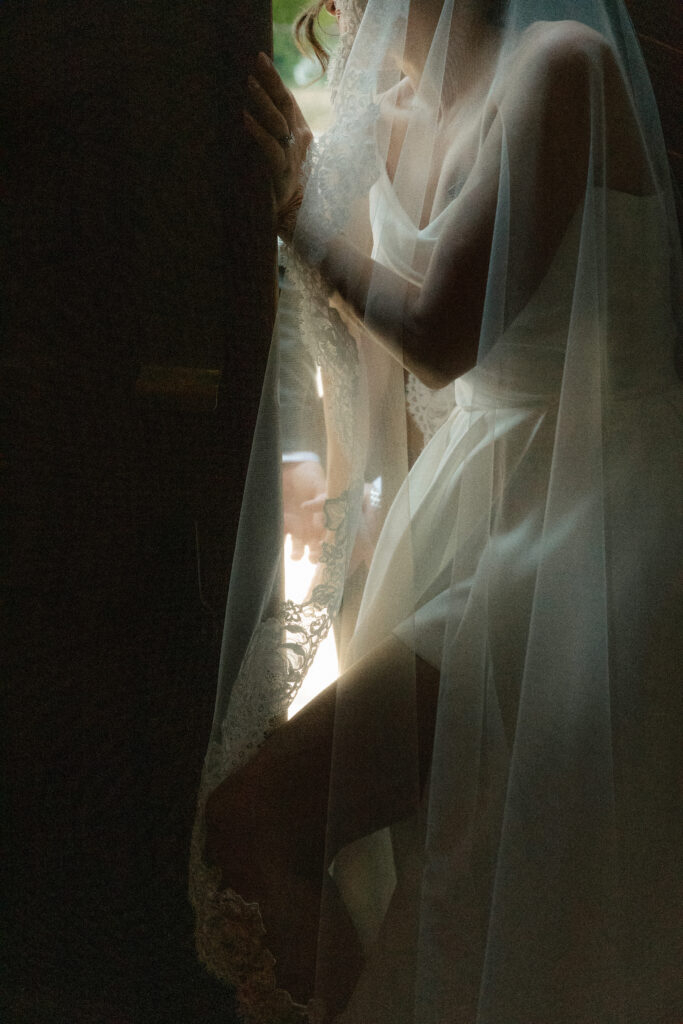 A bride leans out of a cathedral door with light cascading over the pleats of her wedding dress and through her veil while the rest of her is in shadow.