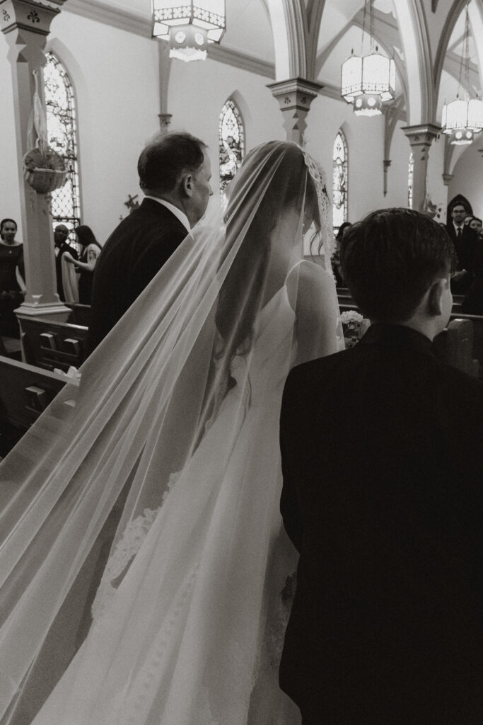 A bride's veil and train trail behind her while her dad and son escort her down the aisle, in black and white.