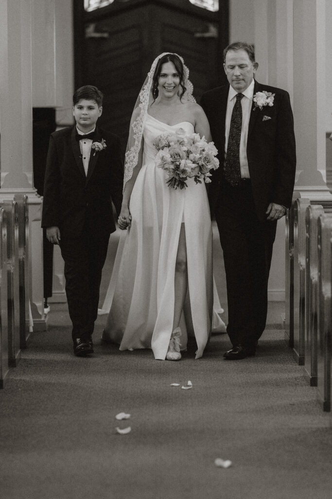 A bride is walked down the aisle of a catholic cathedral by her son and father, in black and white.