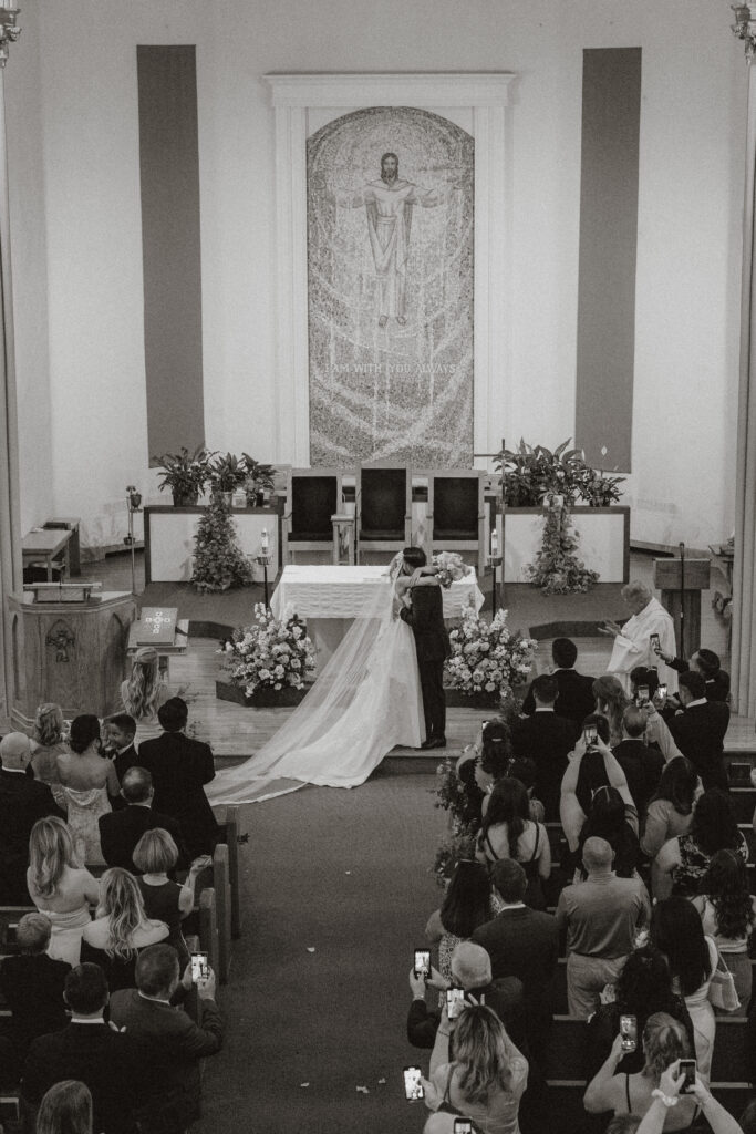 An overhead view of a bride and groom sharing their first kiss as husband and wife in a cathedral, in black and white.