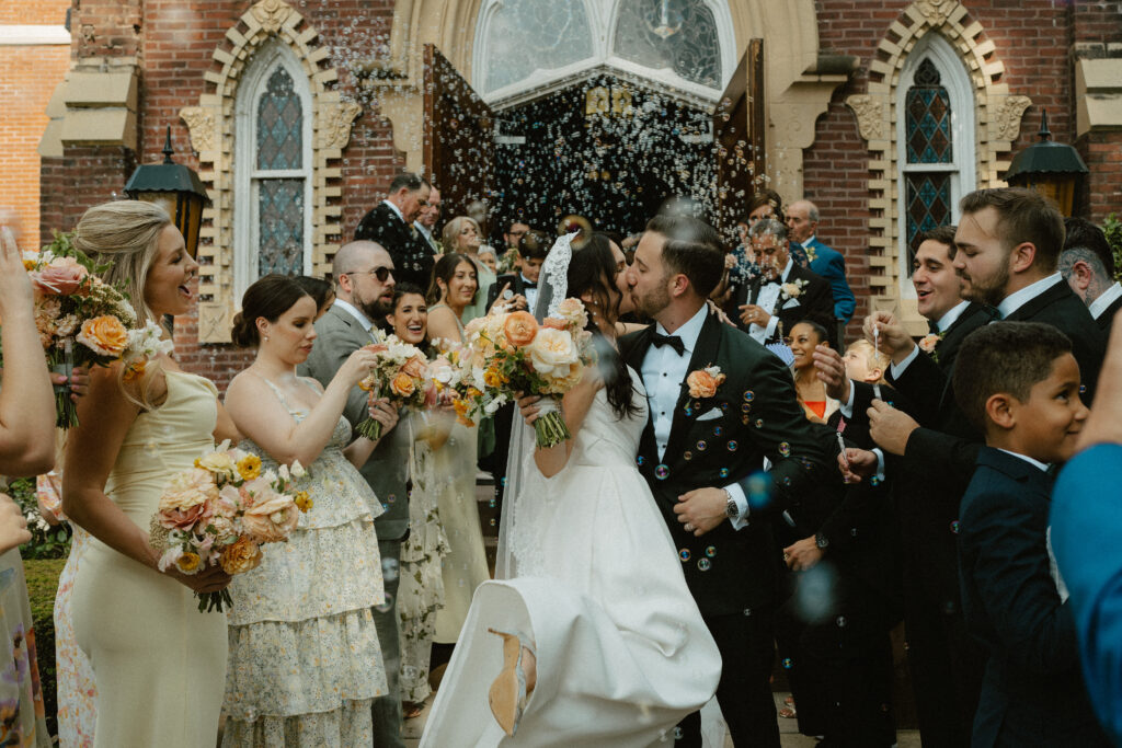 A bride and groom share a kiss after exiting the cathedral ceremony with their loved ones blowing bubbles all around them.