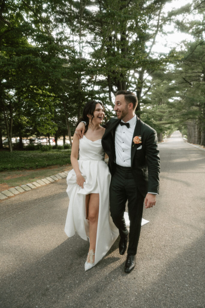 A bride and groom walk arms around each other down a path covered by green trees.