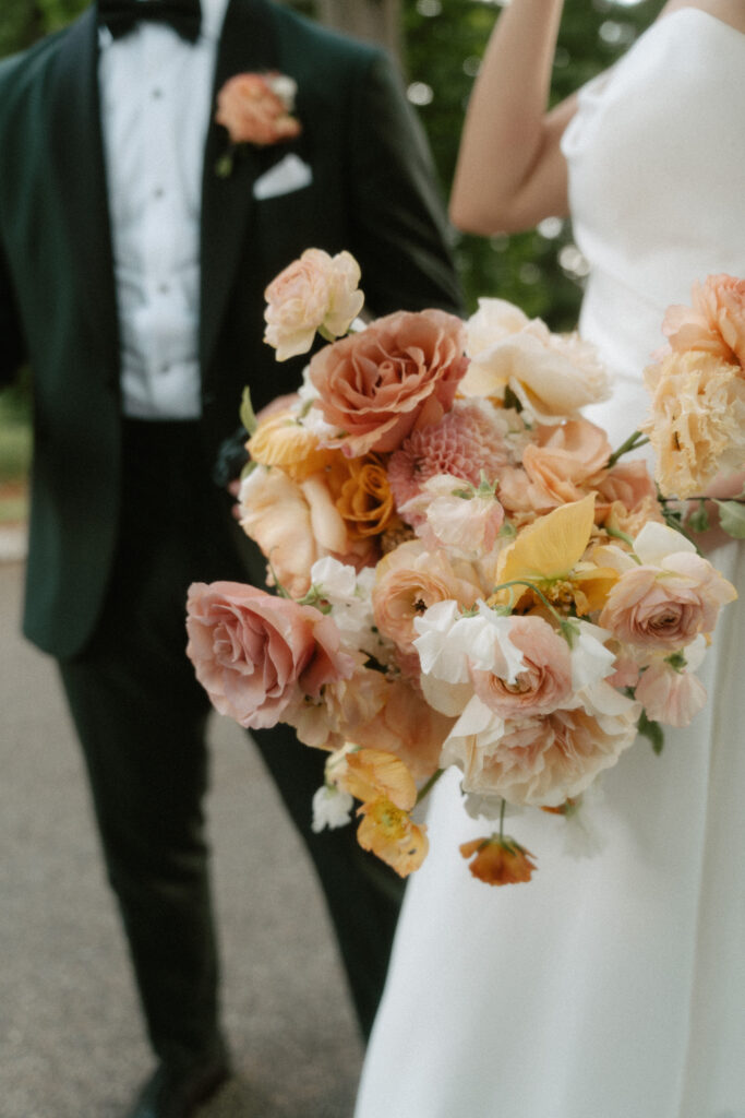 Casually holding her bouquet, a bride shows off her summer bouquet with shades of peach, yellow, and greenery.