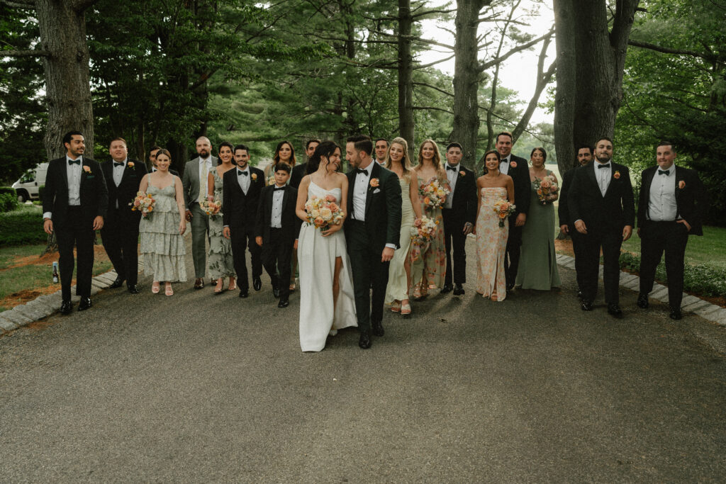 A wedding party in black tuxes and shades of summer greens walk along a path under a canopy of green trees.