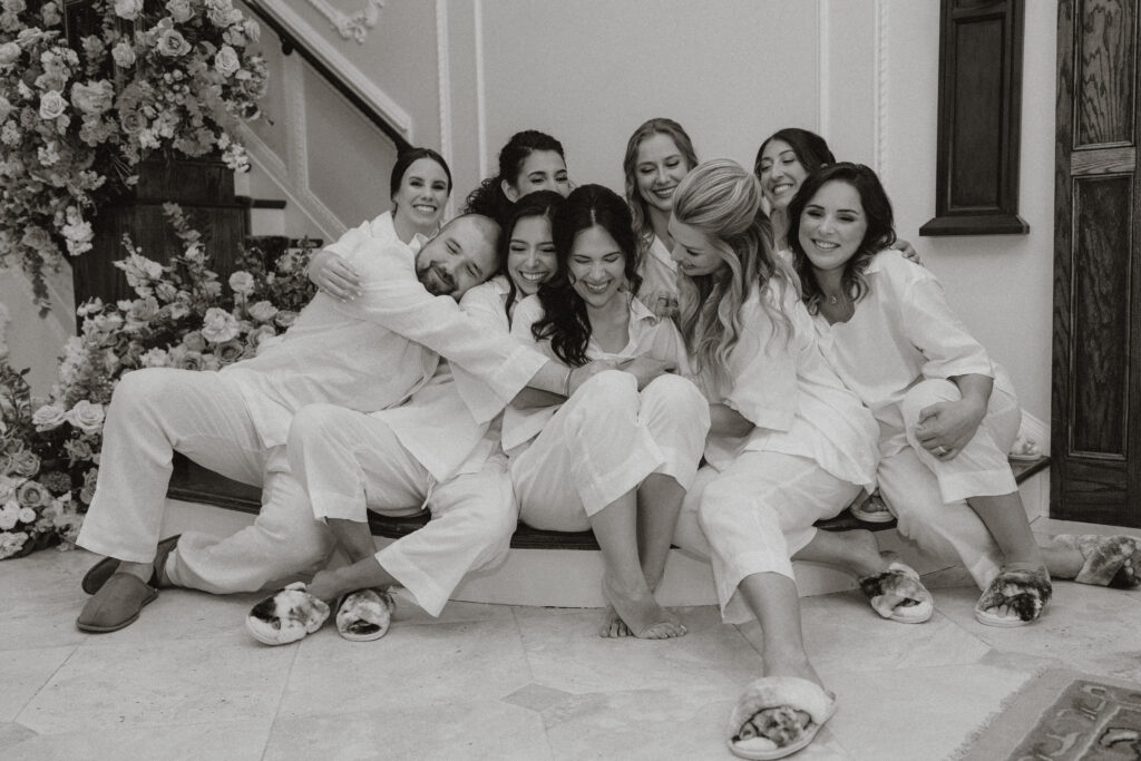 In black and white, a bride laughs with her wedding party in their matching pajamas.