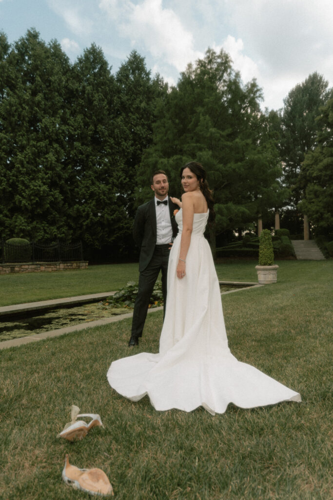 A bride and groom pose together with her shoes kicked off in the grass in a European style garden with a lily pond.