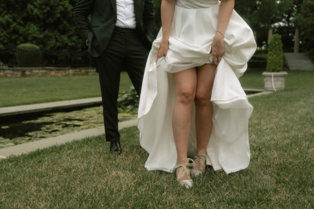 A bride lifts the hem of her wedding dress to show off her blue floral heels while walking through a garden with her husband.