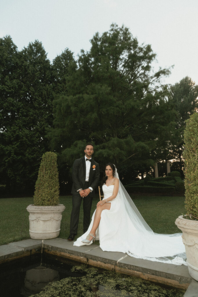 While in a lush garden, a bride sits in a chair with her train and veil flowing around her while her groom stands handsomely beside her.