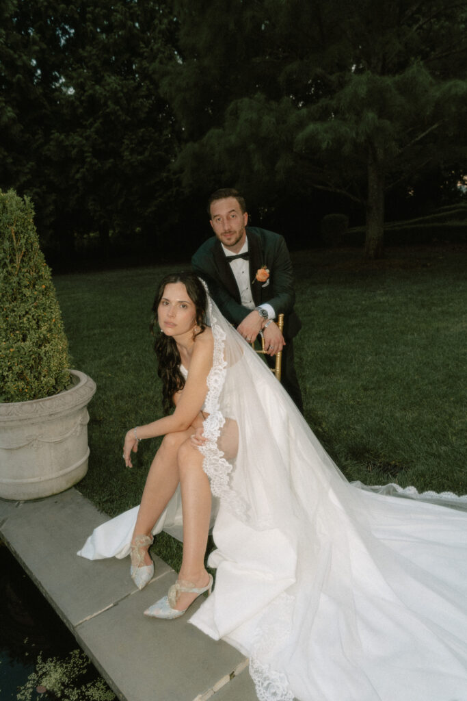 An editorial, direct flash portrait of a bride leaning on her knees while sitting on a chair in a luxurious garden with her groom leaning on the back of the chair.