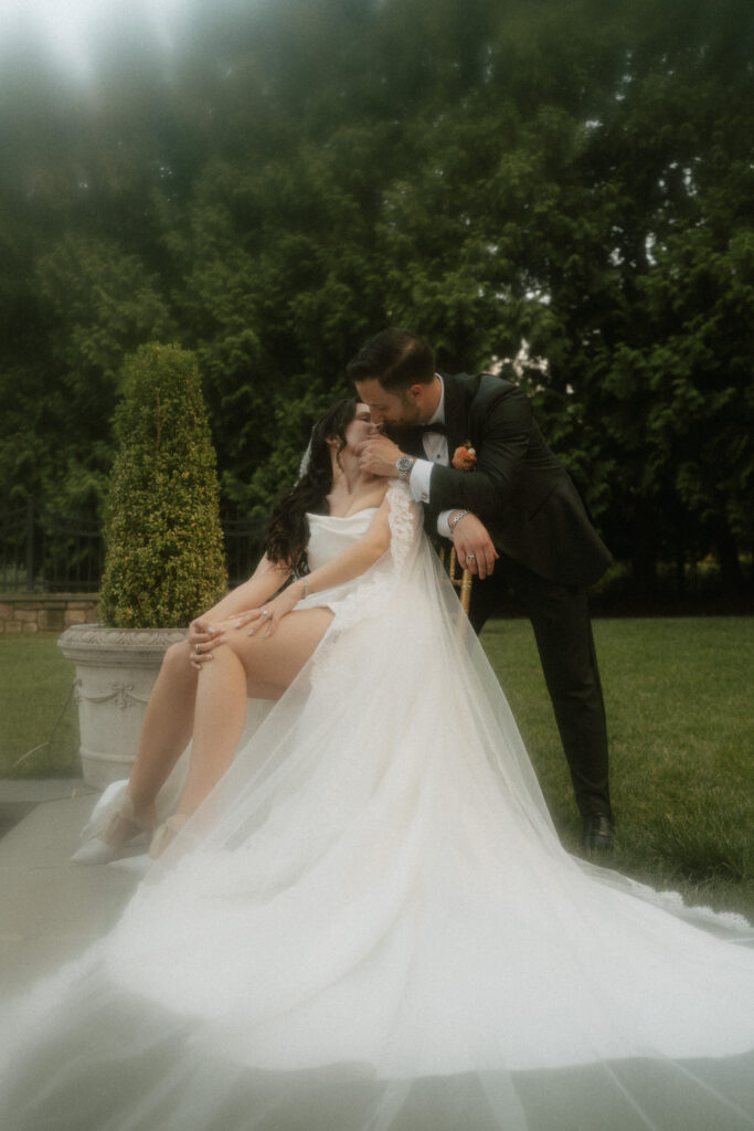 A dreamy and romantic moment between a bride sitting in a chair and leaning back to kiss her groom while in a luxurious garden at their wedding venue.