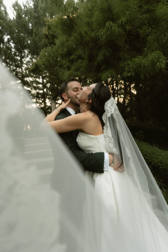 In a luxurious garden, a bride hugs her groom with a big laugh of excitement and her veil flying into the foreground.