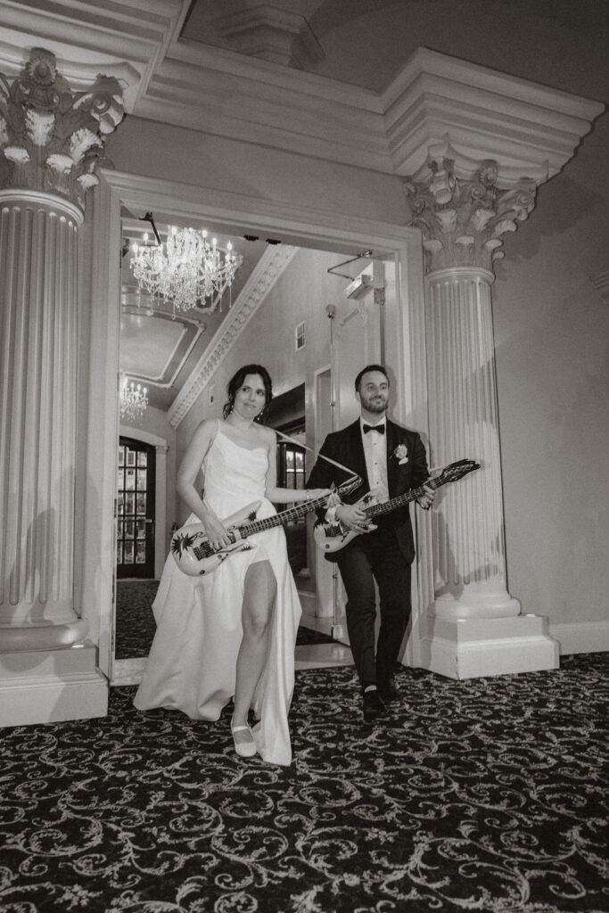 A bride and groom enter their wedding reception like rockstars pretending to play on inflatable guitars, in black and white.