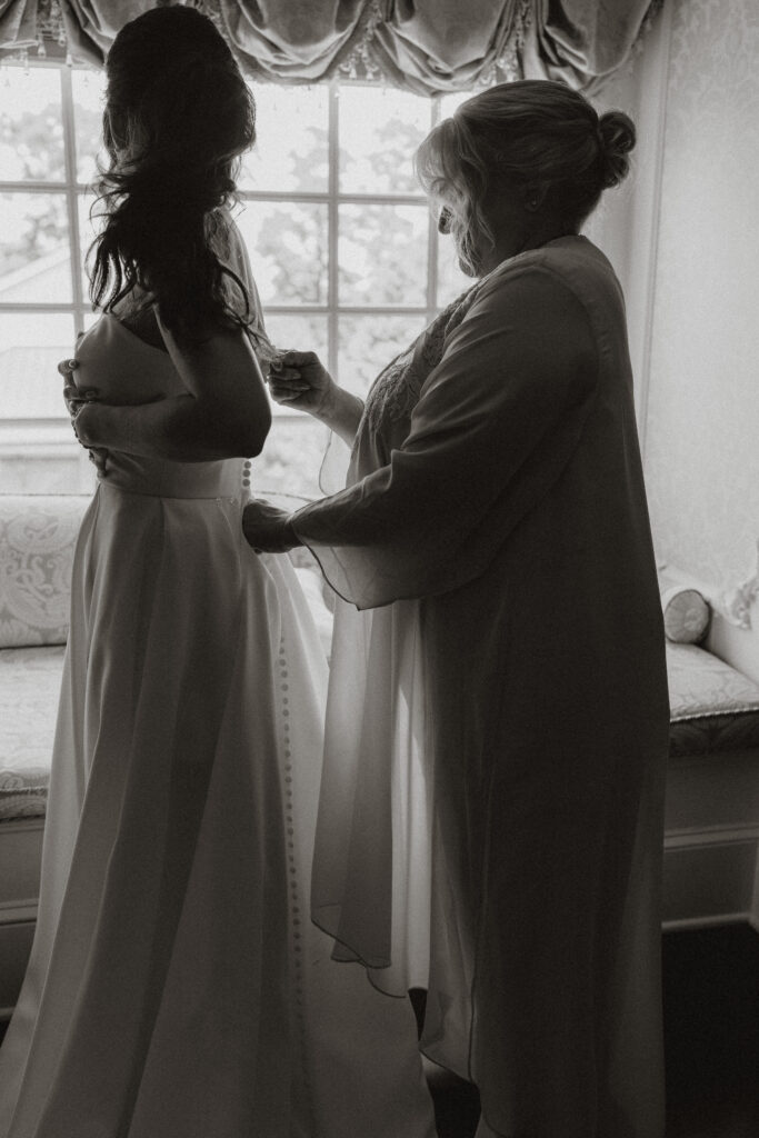 While standing silhouetted in a window, a bride's mother helps her button her wedding dress.