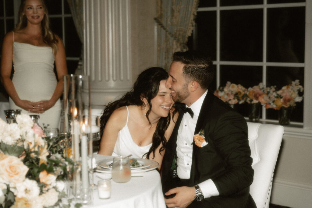 With luxurious florals surrounding them, a bride leans into her groom at their sweetheart table and share a laugh during toasts.