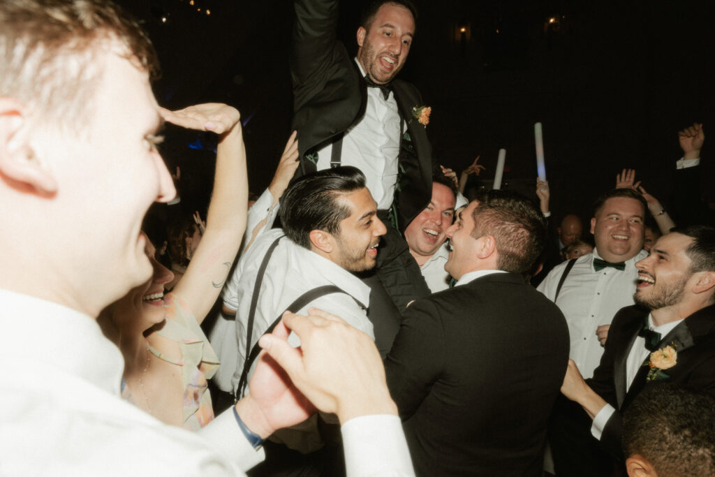 A groom is lifted up in the air while on a chair during the dancing at his wedding reception.