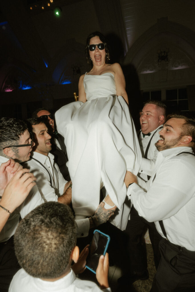 A bride is lifted up in the air while on a chair during the dancing at her wedding reception.