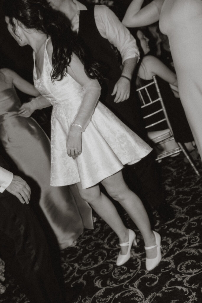 In black and white, a close up of a Bride's second outfit- a short satin a-line dress with sheer gloves and white pumps.