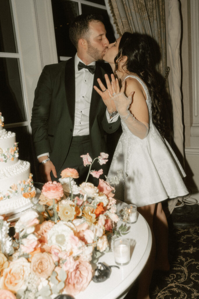 A bride and groom show off their new wedding rings while kissing after their cake cutting.