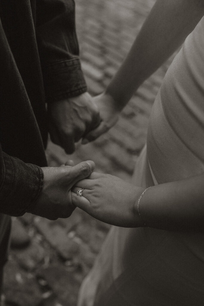 Black and white overhead image of couple's hands clasped showing off engagement ring with cobble stone streets bringing texture to photo