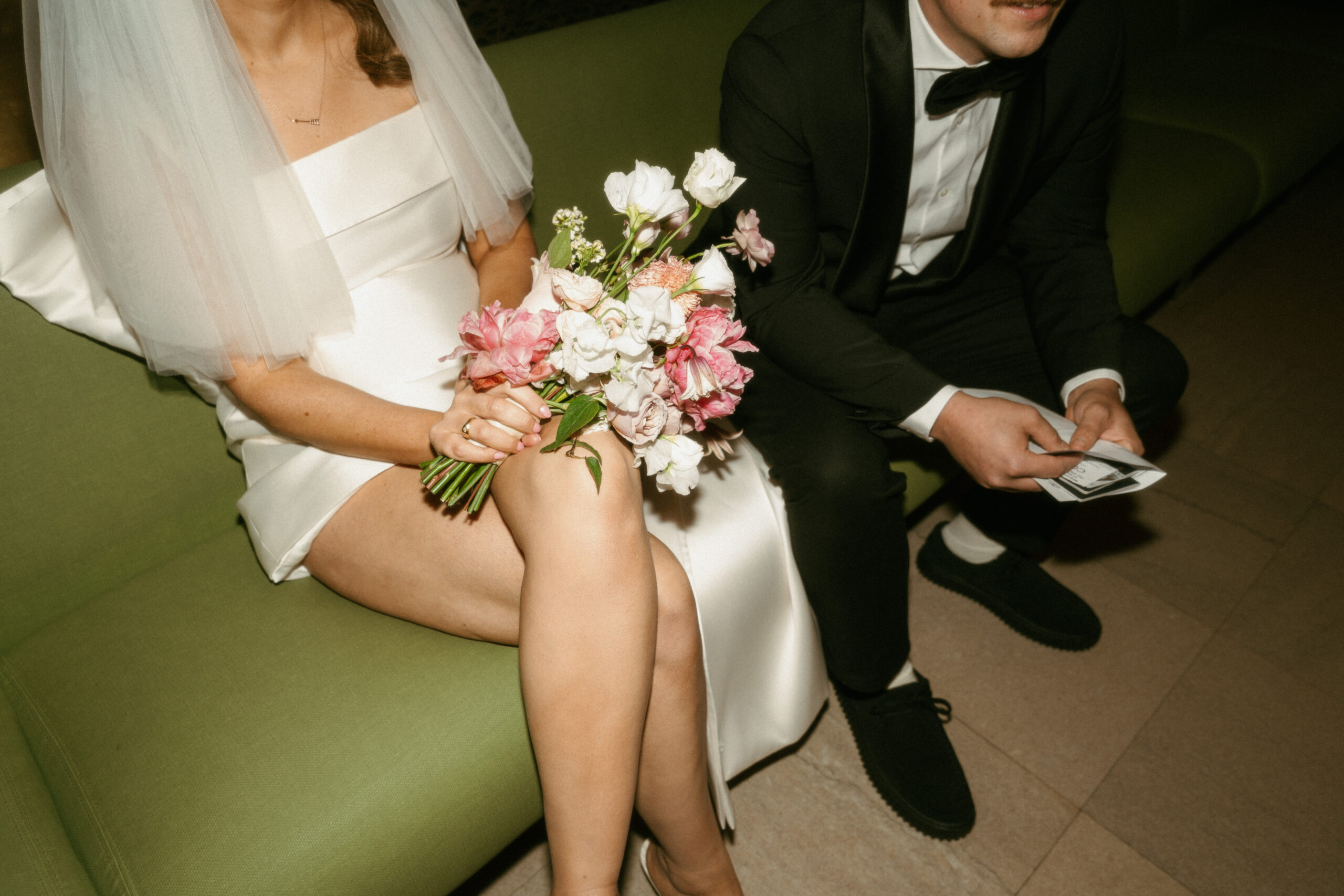 A bride holds her white and pink bouquet on her lap while sitting next to her groom on a green couch.