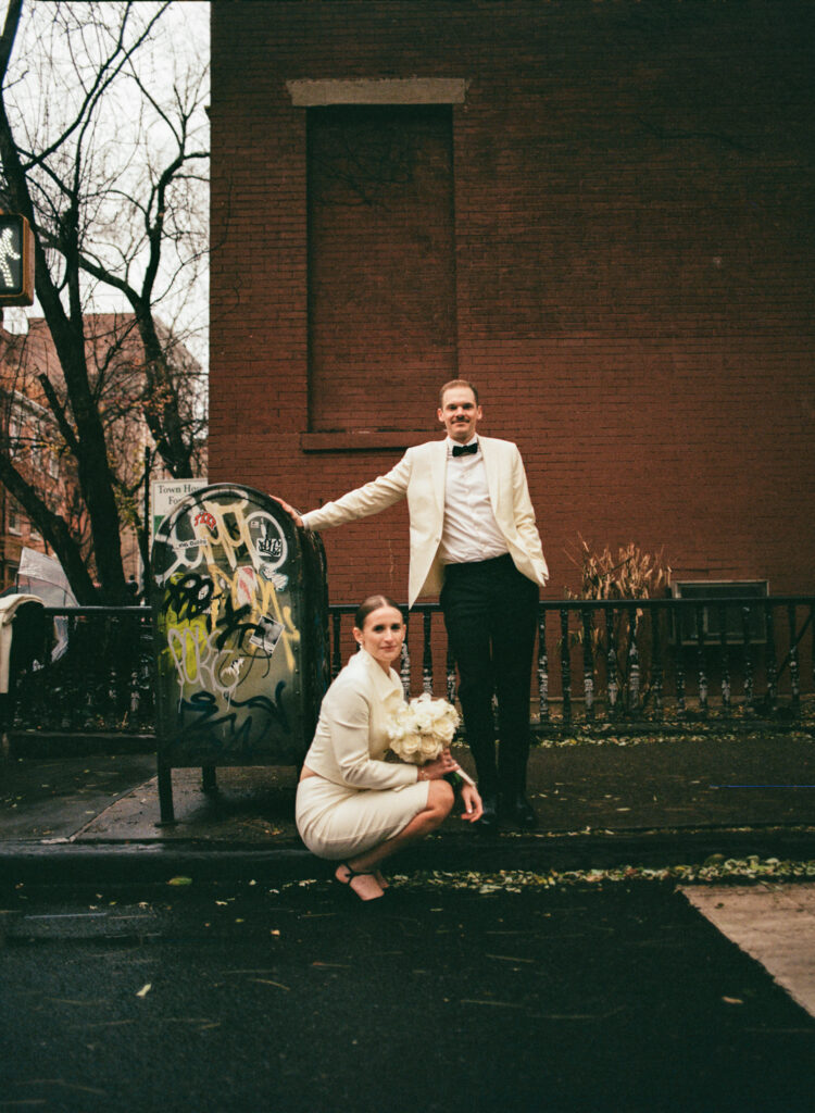 bride crouched down in street with groom leaning on a mailbox with graffiti 