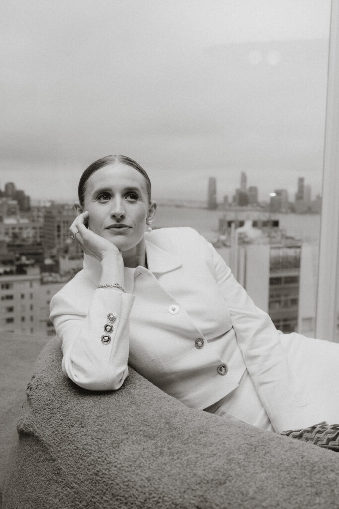 Black and White image of Bride looking up resting chin on palm in front of window with cityscape of NY in the background