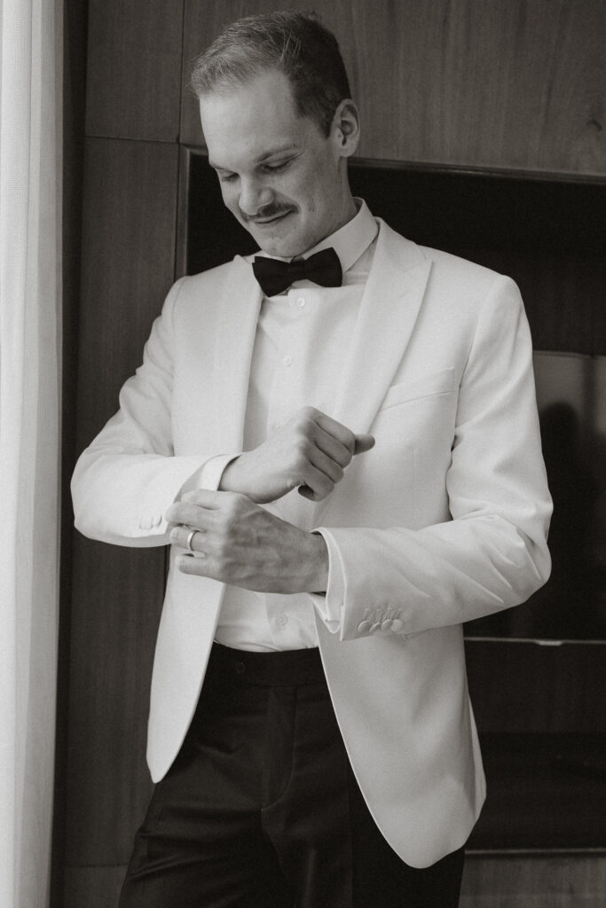 Groom adjusting cuffs on off white jacket with white dress shirt and black bow tie