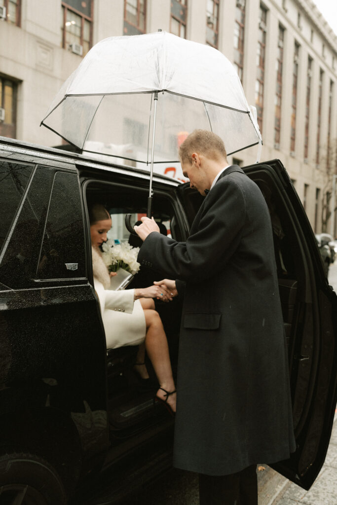 Groom holding clear umbrella for bride to exit SUV at NY City Hall