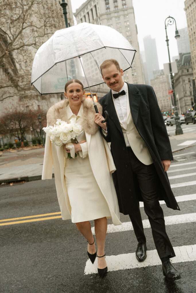 Rainy NYC street crossing with Bride and Groom walking toward camera under clear umbrella