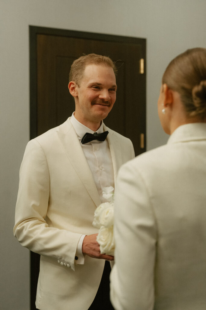 front facing groom smiling at bride in off white suit jacket and black bow tie