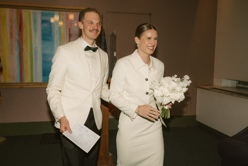 Exiting city hall with marriage license in grooms hand and white rose bouquet in brides hand