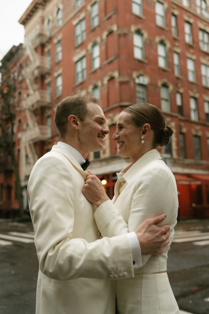 Intimate exchange of grins with couple using apartment building for background to their off white suits