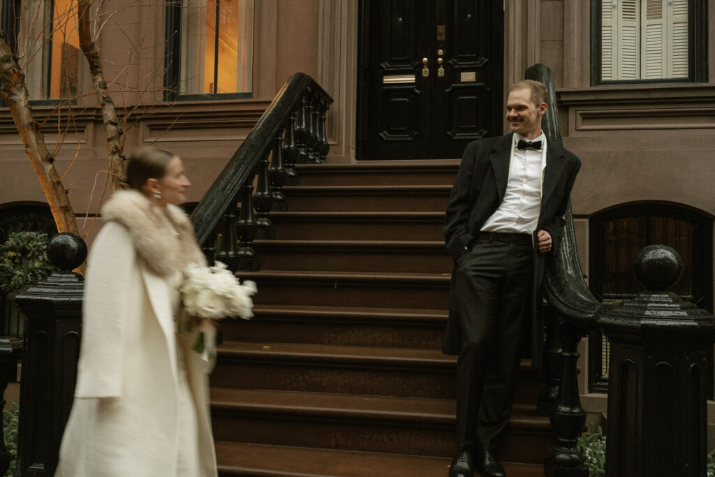 Groom leaning on stair rail of brownstones watching his bride approach